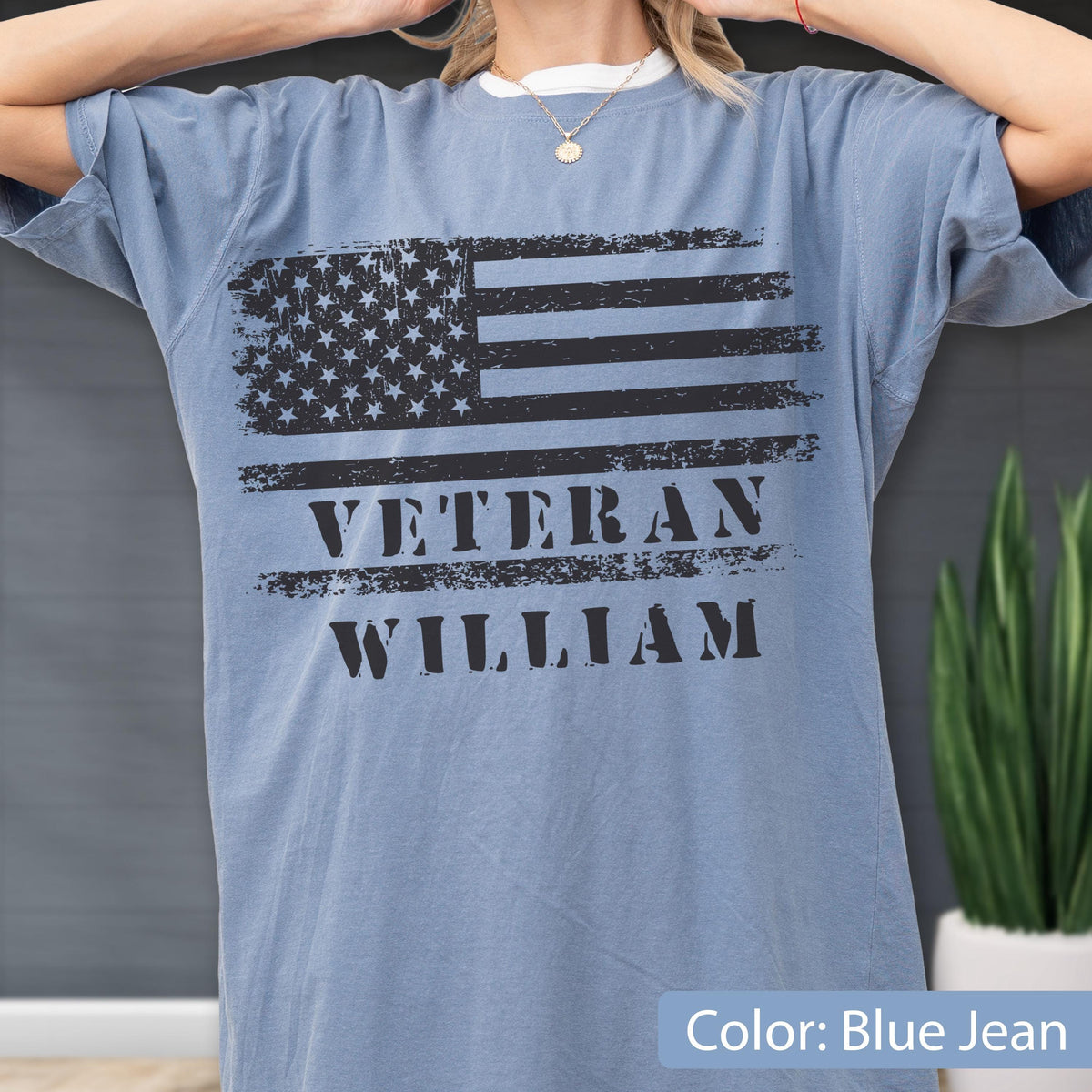 May include: A blue jean colored short-sleeve t-shirt featuring a distressed black graphic of the American flag. Below the flag, the text VETERAN WILLIAM is printed in a bold, distressed black font. The shirt has a crew neckline and loose fit.