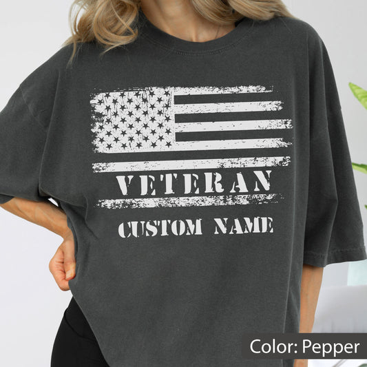 May include: A charcoal grey t-shirt featuring a distressed white graphic of the American flag. Below the flag, the word VETERAN is printed in a bold, distressed white font, followed by the text CUSTOM NAME in the same style and color.