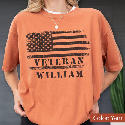 May include: A yam-colored short-sleeve t-shirt featuring a distressed black graphic of the American flag. Below the flag, the text reads VETERAN WILLIAM in a bold, distressed black font. The shirt has a crew neckline and a relaxed fit.