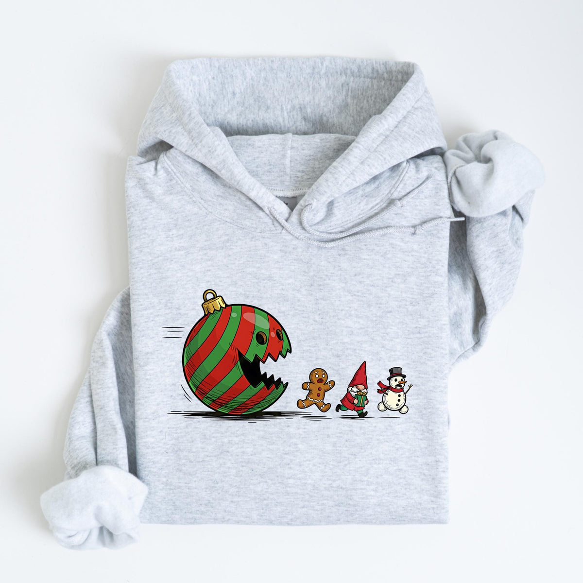 May include: A light gray hoodie featuring a whimsical Christmas-themed graphic. The design depicts a cartoon ornament with a bite taken out of it, chasing a gingerbread man, a gnome, and a snowman. The ornament is red and green striped.