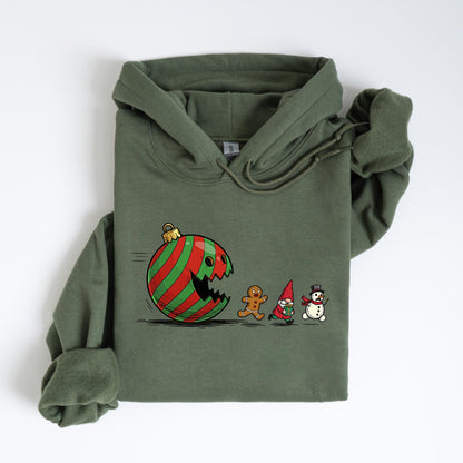 May include: An olive green hoodie featuring a whimsical Christmas-themed graphic. The design depicts a red and green striped ornament with a gaping mouth chasing a gingerbread man, a gnome, and a snowman. The hoodie has a drawstring hood and a kangaroo pocket.