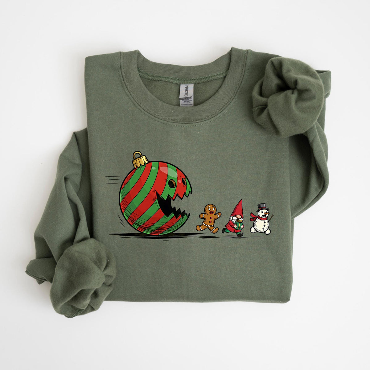 May include: Olive green sweatshirt featuring a humorous Christmas-themed graphic. The design depicts a red and green striped ornament with a bite taken out of it, chasing a gingerbread man, a gnome, and a snowman. The sweatshirt has long sleeves and a crew neck.