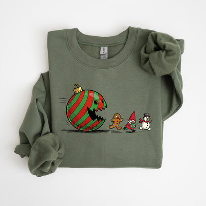 May include: Olive green sweatshirt featuring a humorous Christmas-themed graphic. The design depicts a red and green striped ornament with a bite taken out of it, chasing a gingerbread man, a gnome, and a snowman. The sweatshirt has long sleeves and a crew neck.