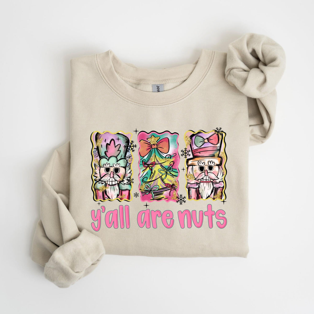 May include: Beige sweatshirt with a colorful graphic of three nutcrackers and the text "y'all are nuts" in pink. The nutcrackers are in a watercolor style with pink, green, yellow, and blue accents. The sweatshirt has a crew neck and long sleeves.