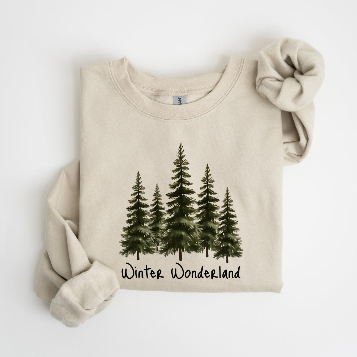 May include: A beige sweatshirt with a watercolor illustration of five evergreen trees. The words "Winter Wonderland" are printed in black script below the trees. A matching scrunchie rests on the right side.