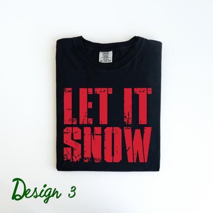 May include: Black t-shirt with the words "LET IT SNOW" in distressed red lettering. The shirt is folded neatly and has a small tag at the neckline. The words "Design 3" are written in green script at the bottom left.