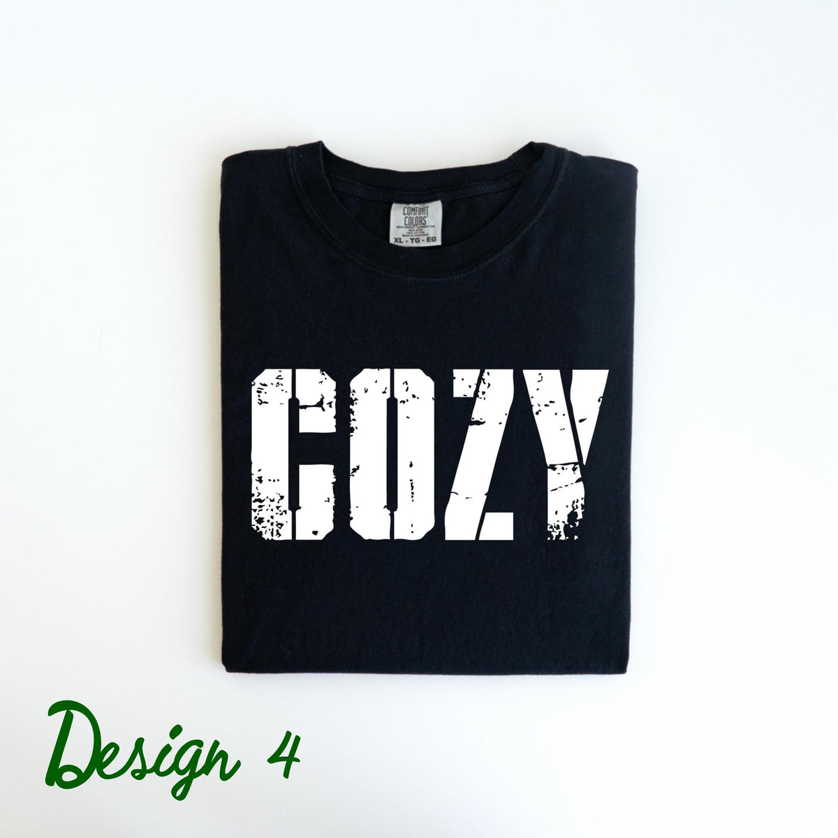 May include: Black t-shirt with the word "COZY" printed in distressed white lettering. The shirt is folded neatly and has a tag at the neckline. The words "Design 4" are written in green script at the bottom.