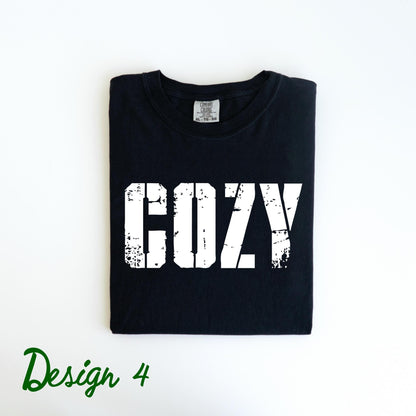 May include: Black t-shirt with the word "COZY" printed in distressed white lettering. The shirt is folded neatly and has a tag at the neckline. The words "Design 4" are written in green script at the bottom.