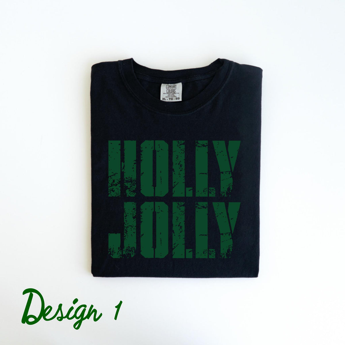 May include: Black t-shirt with the words "HOLLY JOLLY" in distressed green lettering. The shirt is folded neatly on a white surface. The words "Design 1" are written in green script at the bottom left.