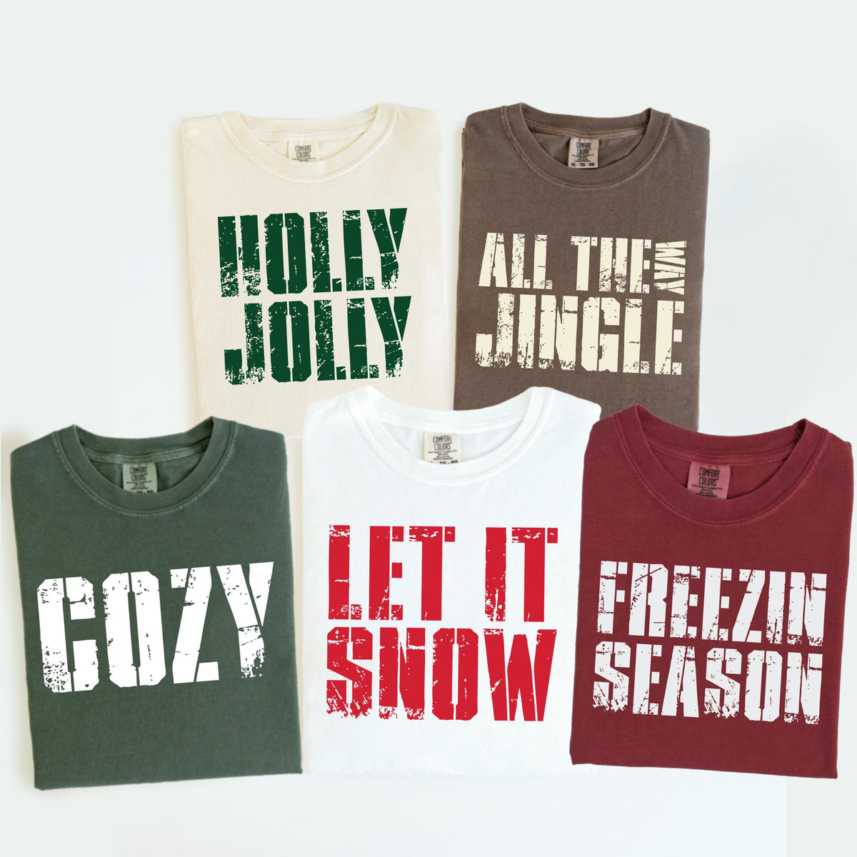 May include: Five t-shirts with holiday-themed text. The shirts are cream, brown, dark green, white, and burgundy. Text includes "HOLLY JOLLY", "ALL THE JINGLE WAY", "COZY", "LET IT SNOW", and "FREEZIN SEASON" in distressed white lettering.