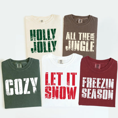 May include: Five t-shirts with holiday-themed text. The shirts are cream, brown, dark green, white, and burgundy. Text includes "HOLLY JOLLY", "ALL THE JINGLE WAY", "COZY", "LET IT SNOW", and "FREEZIN SEASON" in distressed white lettering.