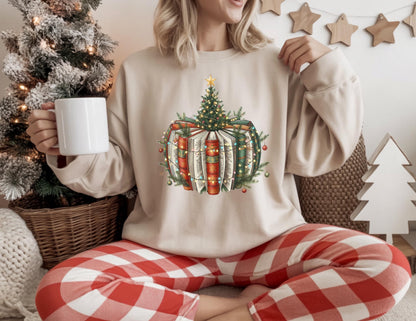 May include: Beige sweatshirt featuring a Christmas tree design atop a stack of books, adorned with lights and ornaments. The person is wearing red and white plaid pants and holding a white mug. A Christmas tree and wooden stars are in the background.