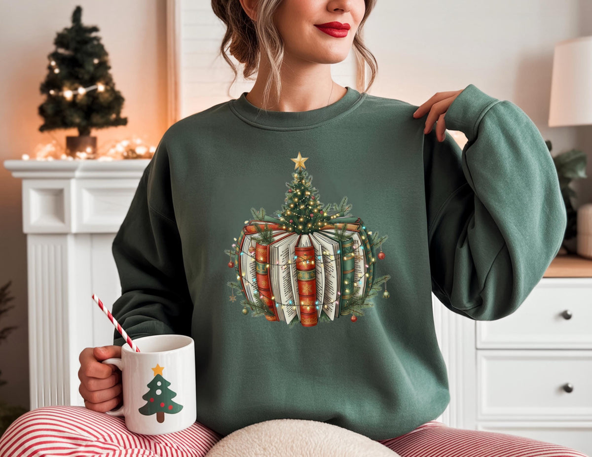 May include: A dark green sweatshirt featuring a Christmas tree design atop a stack of books. The design includes a star, lights, and festive greenery. A white mug with a Christmas tree design is held in the hand.