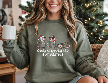 May include: Olive green sweatshirt with the text "OVERSTIMULATED BUT FESTIVE" and illustrations of three opossums wearing a Santa hat, a scarf, and Christmas lights. The sweatshirt is a long-sleeved crew neck.