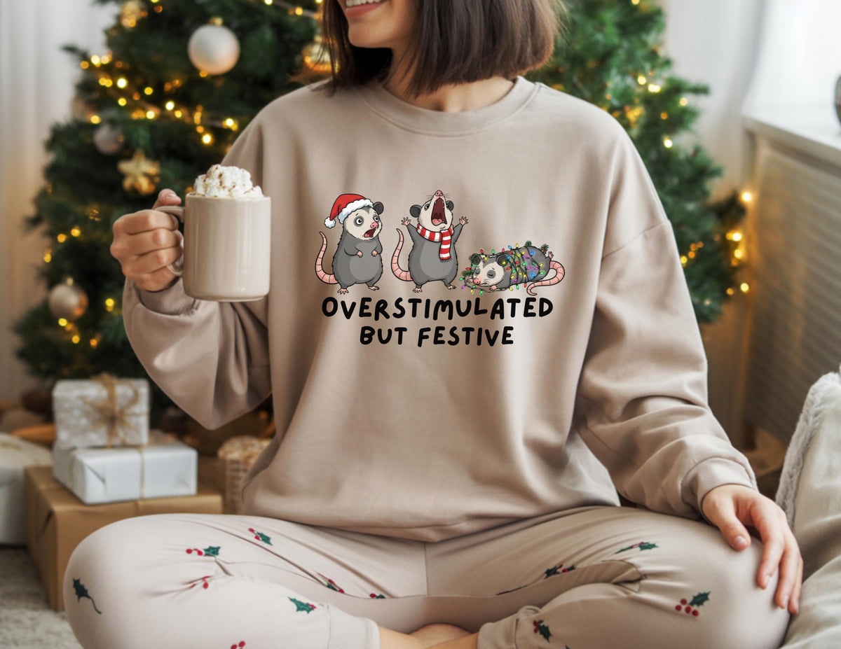 May include: Beige sweatshirt with the text "OVERSTIMULATED BUT FESTIVE" and a graphic of three opossums in festive attire. One opossum wears a Santa hat, another a scarf, and the third is wrapped in Christmas lights. The person is holding a mug.