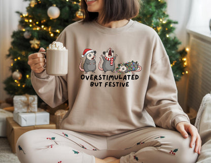 May include: Beige sweatshirt with the text "OVERSTIMULATED BUT FESTIVE" and a graphic of three opossums in festive attire. One opossum wears a Santa hat, another a scarf, and the third is wrapped in Christmas lights. The person is holding a mug.