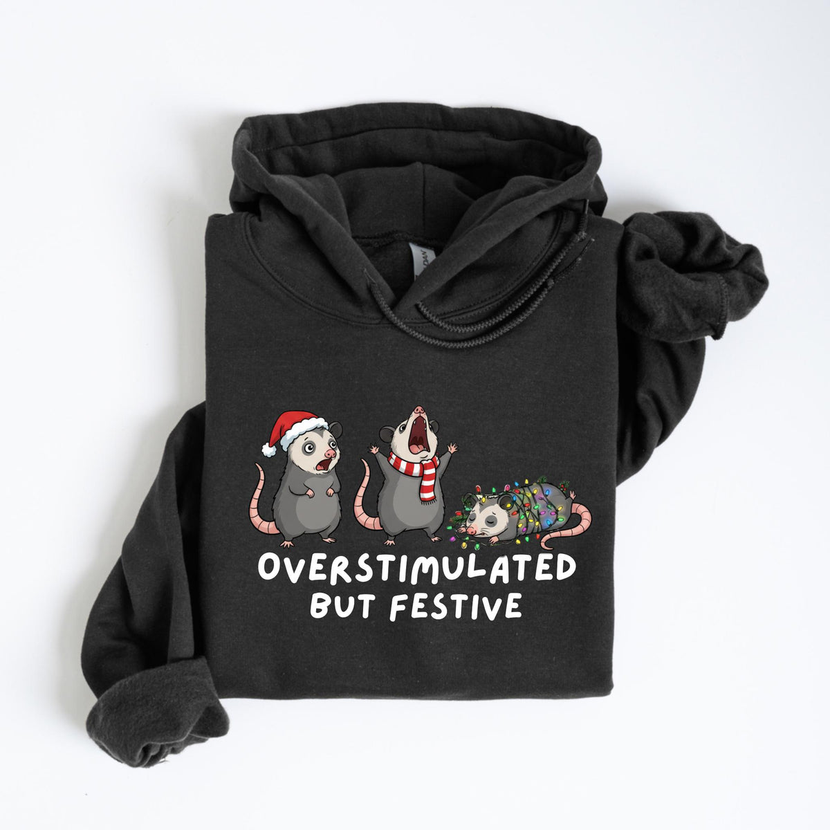 May include: Dark gray hoodie featuring a graphic of three opossums, each adorned with festive holiday accessories. One wears a Santa hat, another a scarf, and the third is wrapped in Christmas lights. The text "OVERSTIMULATED BUT FESTIVE" is printed below.