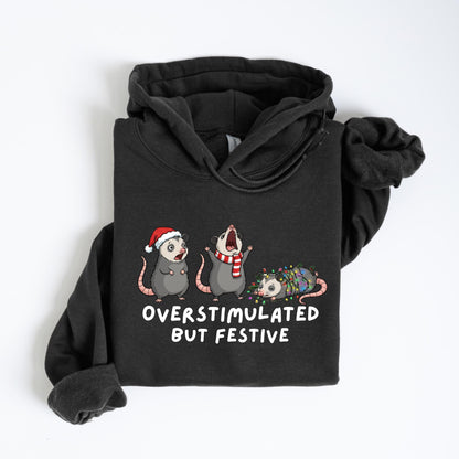 May include: Dark gray hoodie featuring a graphic of three opossums, each adorned with festive holiday accessories. One wears a Santa hat, another a scarf, and the third is wrapped in Christmas lights. The text "OVERSTIMULATED BUT FESTIVE" is printed below.