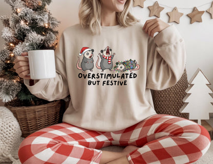 May include: Beige sweatshirt with the text "OVERSTIMULATED BUT FESTIVE" and a graphic of three opossums in holiday attire. The opossums are wearing a Santa hat, a scarf, and Christmas lights. The person is wearing red and white plaid pants.