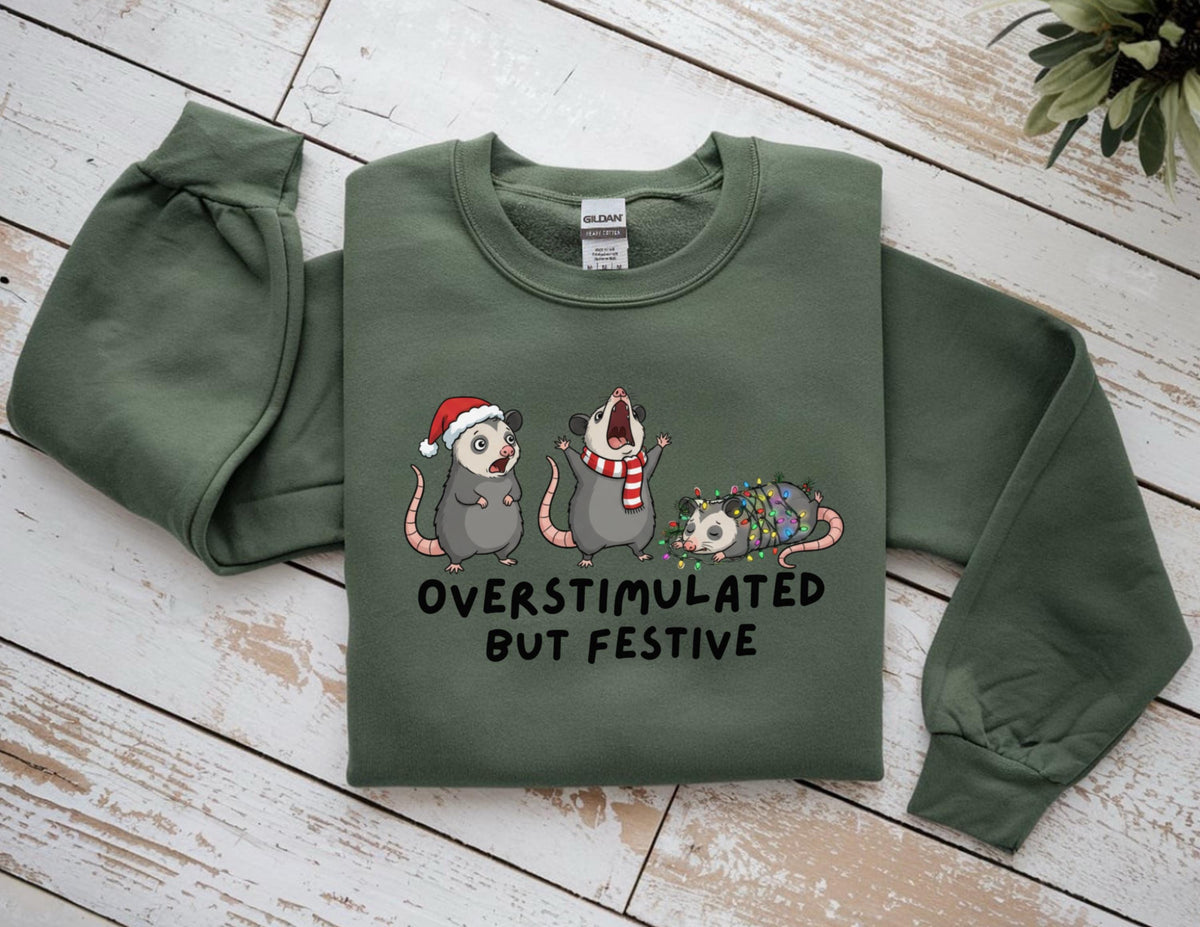 May include: Green sweatshirt with the text "OVERSTIMULATED BUT FESTIVE" and illustrations of three opossums. One opossum wears a Santa hat, another a scarf, and the third is wrapped in Christmas lights. The sweatshirt has long sleeves.