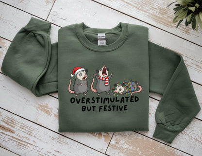 May include: Green sweatshirt with the text "OVERSTIMULATED BUT FESTIVE" and illustrations of three opossums. One opossum wears a Santa hat, another a scarf, and the third is wrapped in Christmas lights. The sweatshirt has long sleeves.
