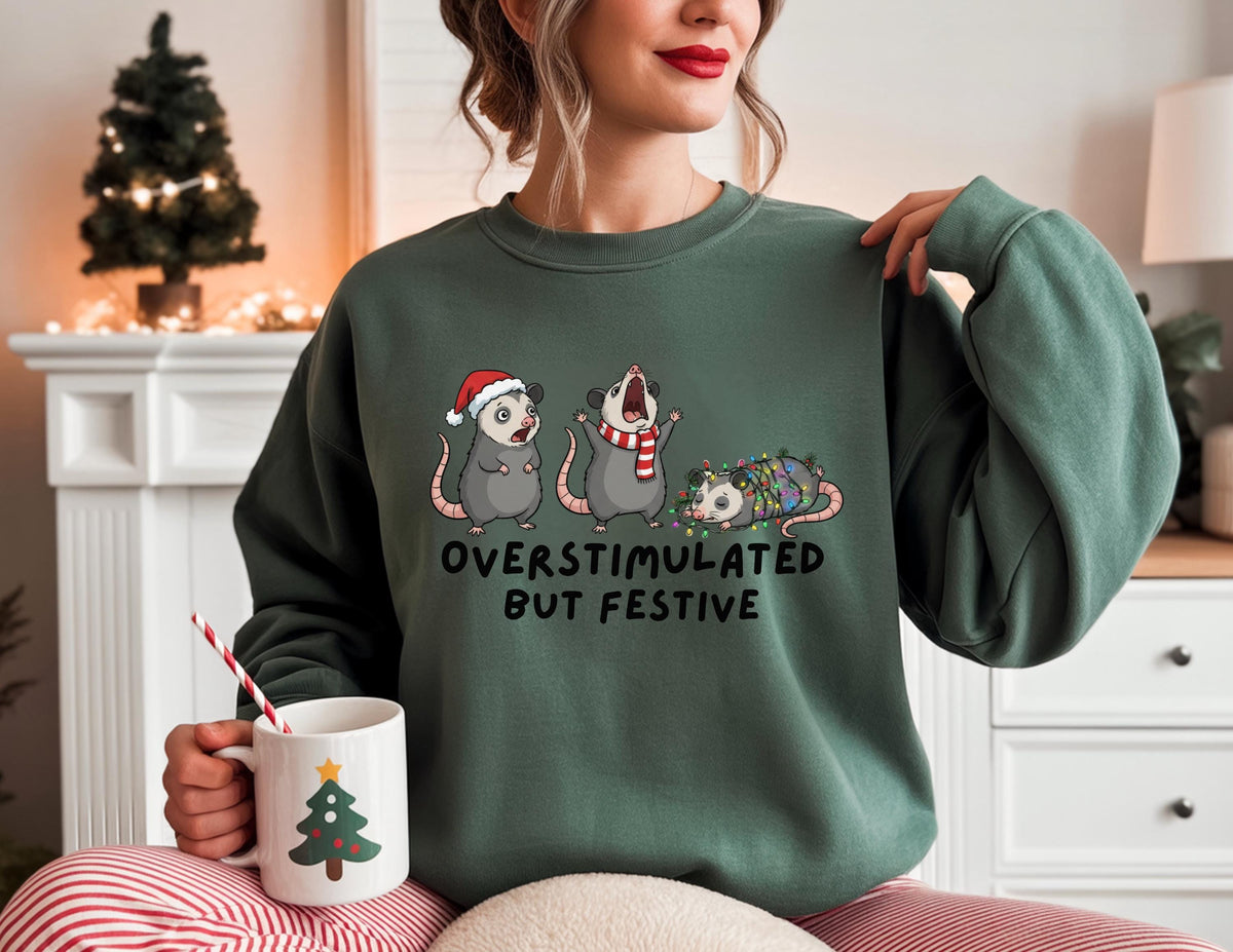 May include: A dark green sweatshirt with the text "OVERSTIMULATED BUT FESTIVE" and a graphic of three opossums, one wearing a Santa hat, one with a scarf, and one wrapped in Christmas lights. A white mug with a Christmas tree design is held in the hand.