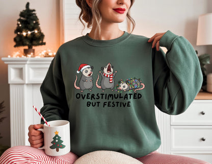 May include: A dark green sweatshirt with the text "OVERSTIMULATED BUT FESTIVE" and a graphic of three opossums, one wearing a Santa hat, one with a scarf, and one wrapped in Christmas lights. A white mug with a Christmas tree design is held in the hand.