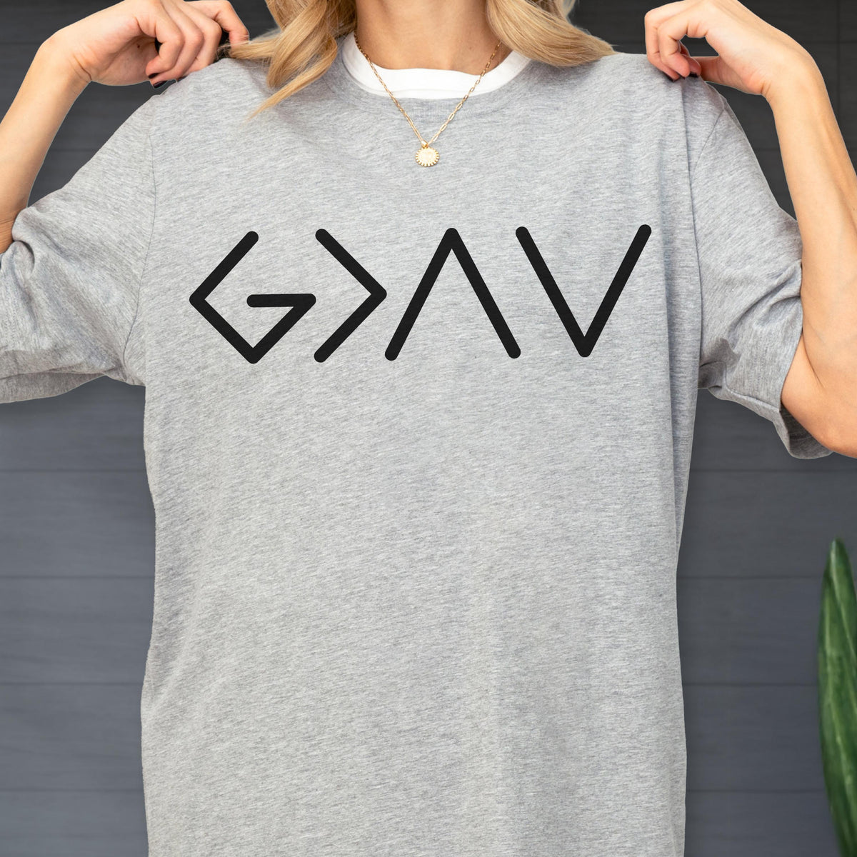 May include: A heather gray short-sleeve t-shirt featuring the black text G > ^ V printed across the chest. The shirt has a crew neckline and a standard fit, showcasing a simple design with a clear graphic print on the front of the garment.