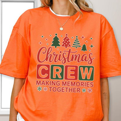 May include: An orange short-sleeved t-shirt featuring a graphic print with four stylized Christmas trees, stars, and the text "Christmas CREW MAKING MEMORIES TOGETHER" in red, green, and white colors, accented with small snowflake icons.
