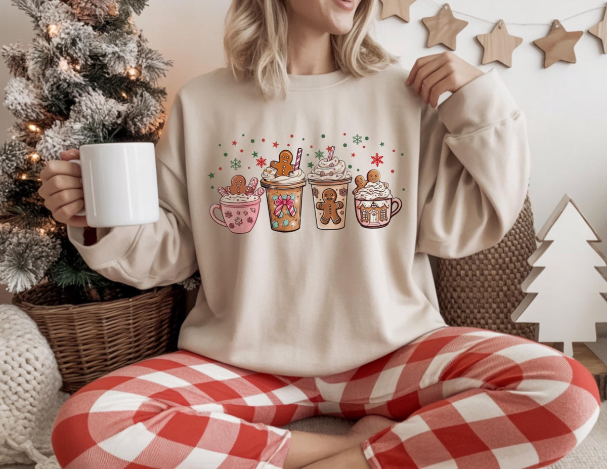 May include: Beige sweatshirt featuring a festive graphic of four holiday-themed coffee cups with gingerbread men and whipped cream. The design includes red, green, and white snowflake and star accents. The person is holding a white mug and wearing red and white plaid pants.