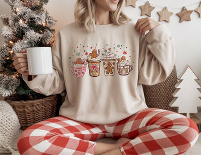 May include: Beige sweatshirt featuring a festive graphic of four holiday-themed coffee cups with gingerbread men and whipped cream. The design includes red, green, and white snowflake and star accents. The person is holding a white mug and wearing red and white plaid pants.