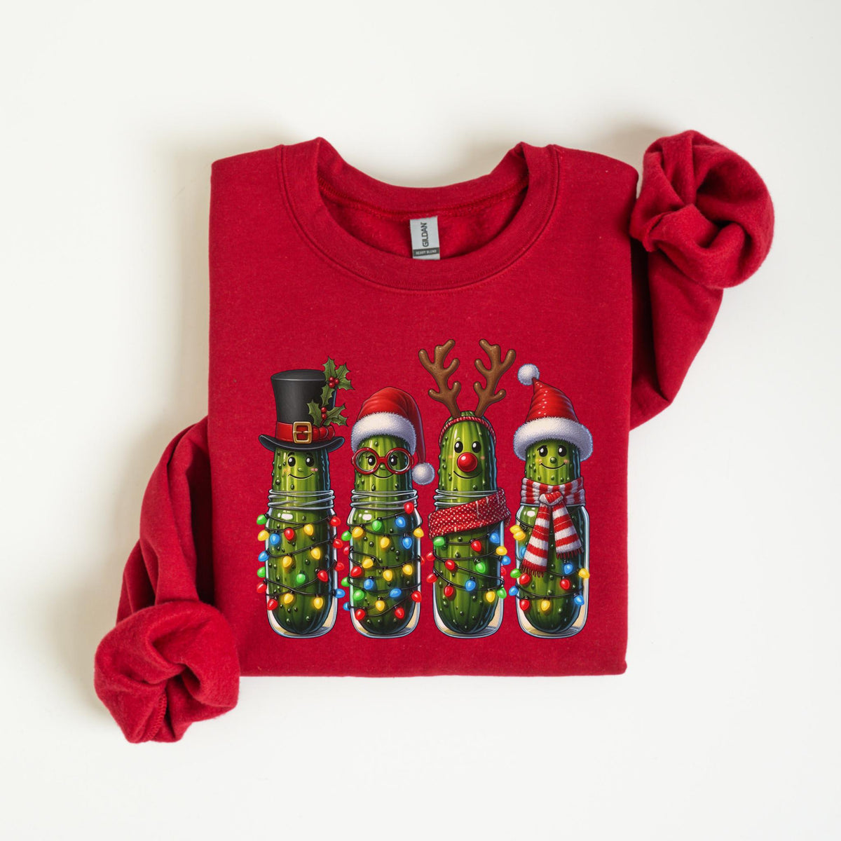 May include: Red crewneck sweatshirt featuring a festive design of four pickles in glass jars, each adorned with Christmas accessories like hats, antlers, and scarves. The pickles are decorated with colorful string lights, creating a holiday theme.