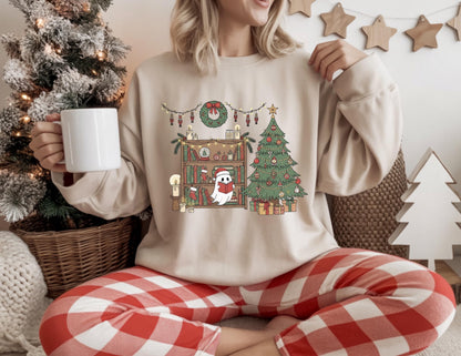 May include: A beige sweatshirt with a Christmas-themed graphic featuring a ghost reading a book in front of a Christmas tree and bookshelf. The ghost wears a Santa hat and scarf. The person is wearing red and white plaid pajama pants and holding a white mug.