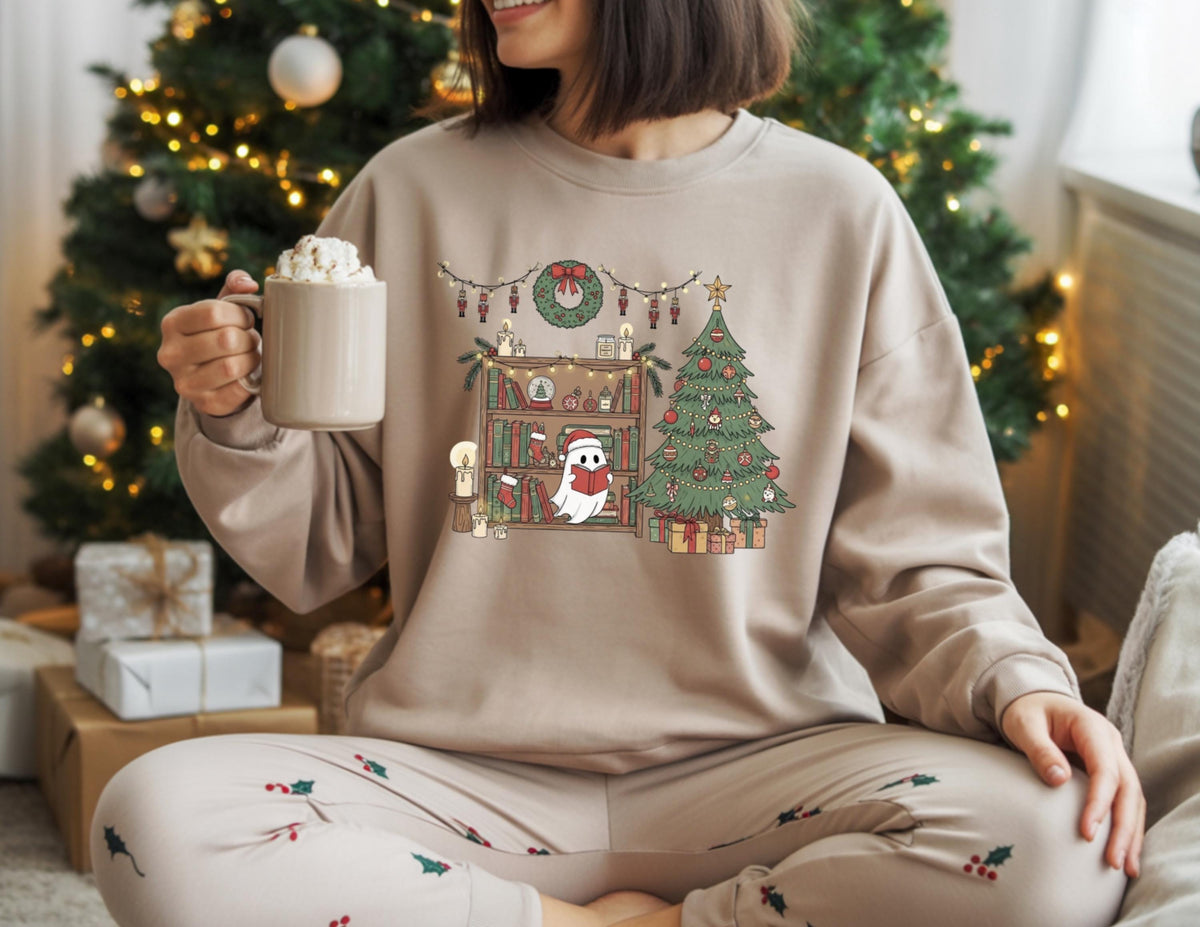 May include: Beige sweatshirt with a Christmas-themed graphic. The graphic features a bookshelf, Christmas tree, and a ghost wearing a Santa hat. The person is holding a mug of whipped cream. The person is wearing matching pajama pants with Christmas holly.