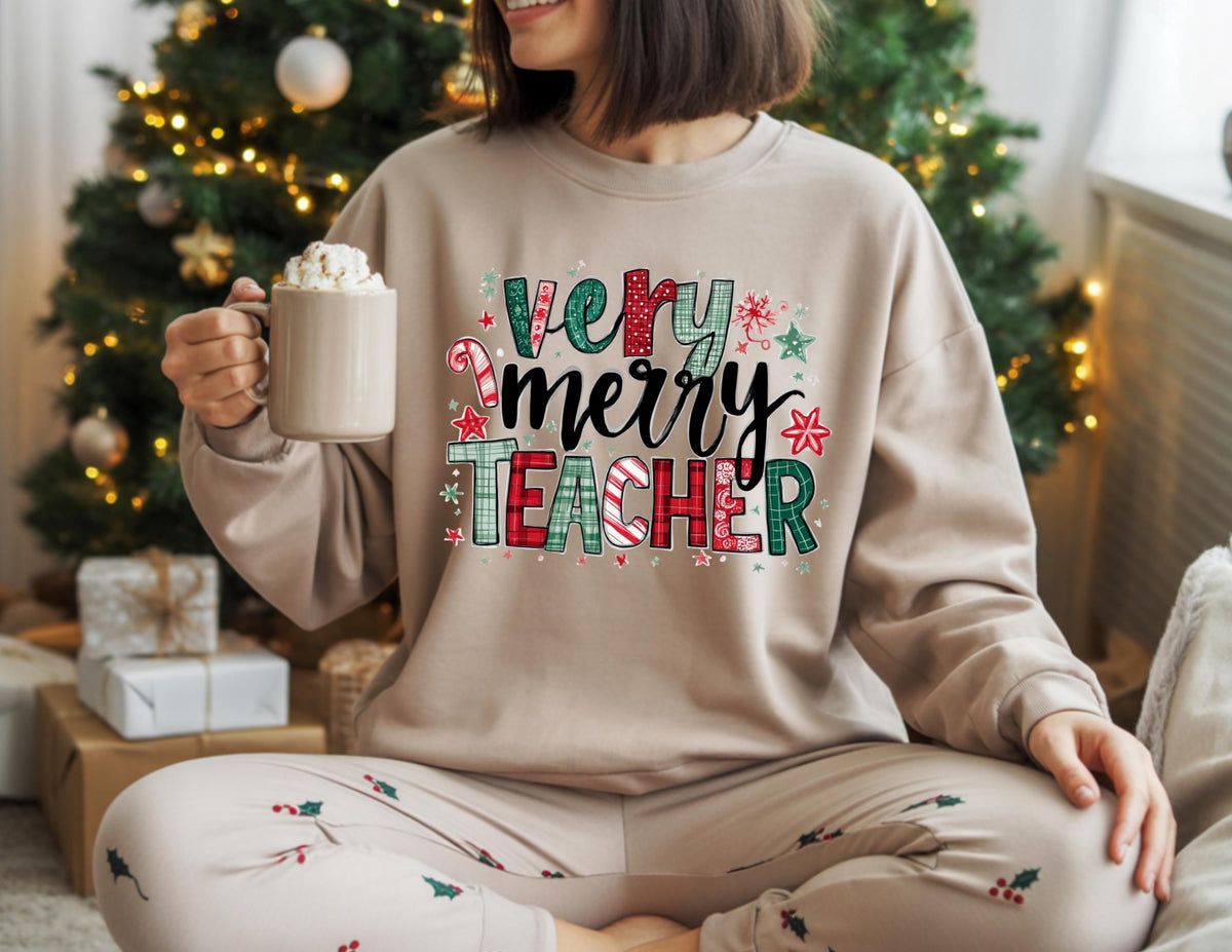 May include: A beige sweatshirt with the text "Very Merry Teacher" in a festive design with red, green, and white accents. The person is holding a mug with whipped cream, and wearing matching Christmas-themed pajamas.