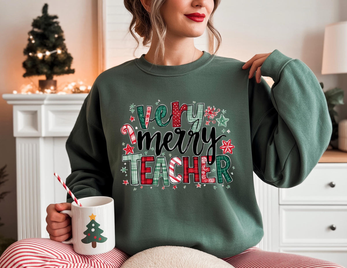 May include: A dark green sweatshirt with the text "Very Merry Teacher" in a festive design. The design includes candy canes, stars, and plaid patterns. A white mug with a Christmas tree design is held in the hand.