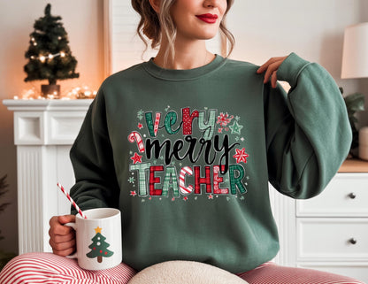 May include: A dark green sweatshirt with the text "Very Merry Teacher" in a festive design. The design includes candy canes, stars, and plaid patterns. A white mug with a Christmas tree design is held in the hand.