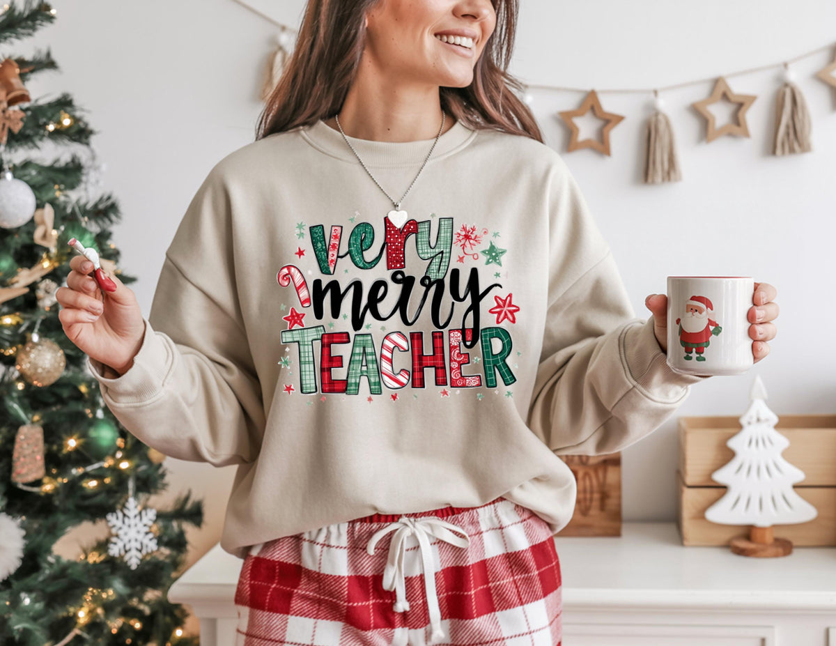 May include: Beige sweatshirt with the text "Very Merry Teacher" in red, green, and black. The sweatshirt is paired with red and white plaid pajama pants. A white mug with a Santa Claus graphic is held in the hand.