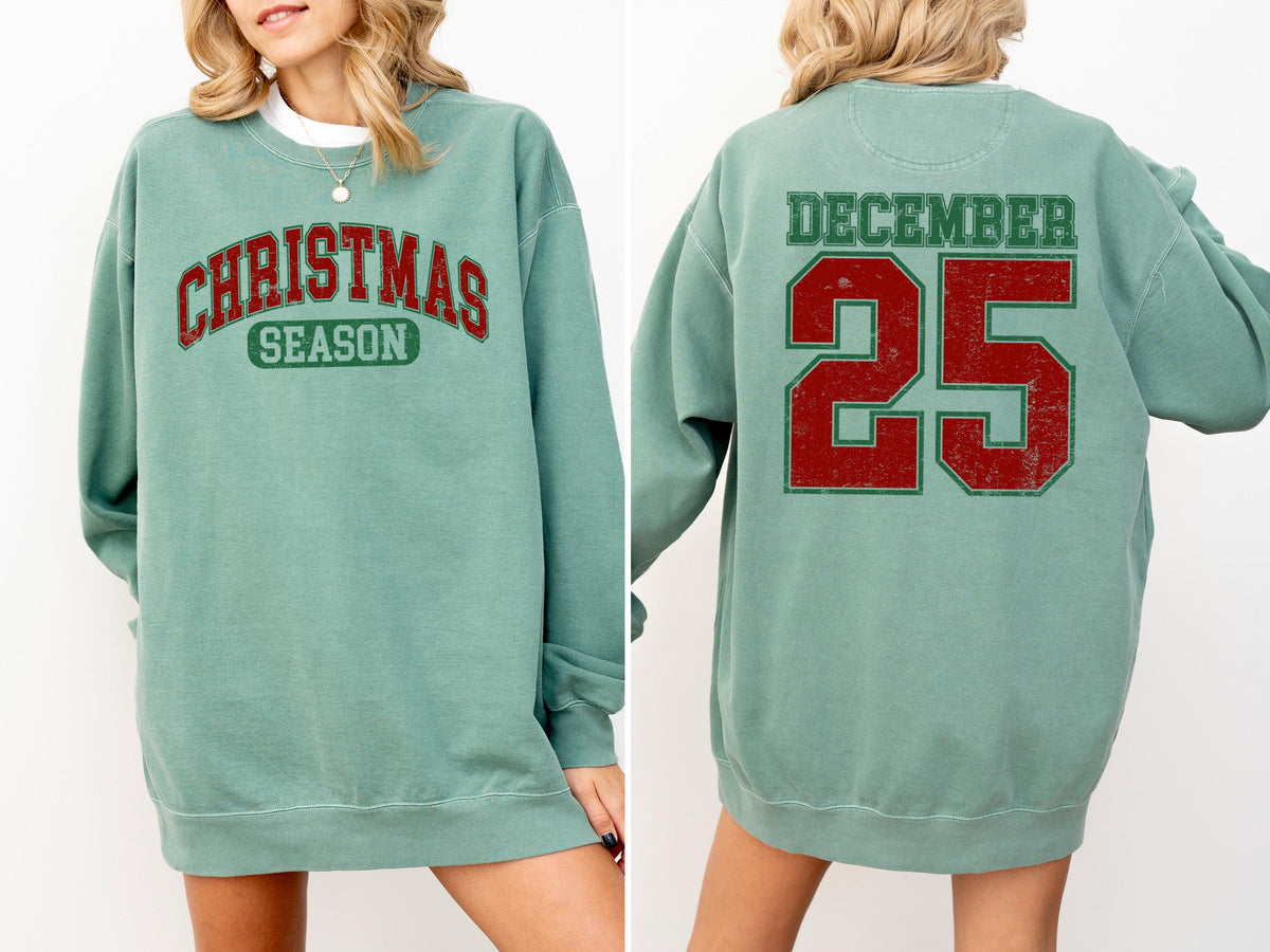 May include: A teal sweatshirt with the words "CHRISTMAS SEASON" in red and green lettering on the front. The back of the sweatshirt displays "DECEMBER 25" in red and green lettering. The sweatshirt is a long-sleeved, crew neck style.