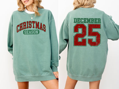 May include: A teal sweatshirt with the words "CHRISTMAS SEASON" in red and green lettering on the front. The back of the sweatshirt displays "DECEMBER 25" in red and green lettering. The sweatshirt is a long-sleeved, crew neck style.