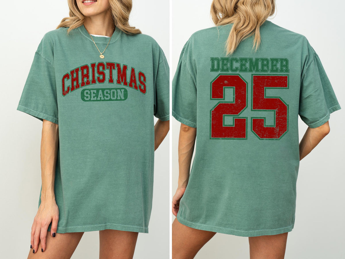 May include: A sage green t-shirt with the words "CHRISTMAS SEASON" in red and green lettering on the front. The back of the shirt displays "DECEMBER 25" in red and green lettering. The shirt is a casual, oversized fit.