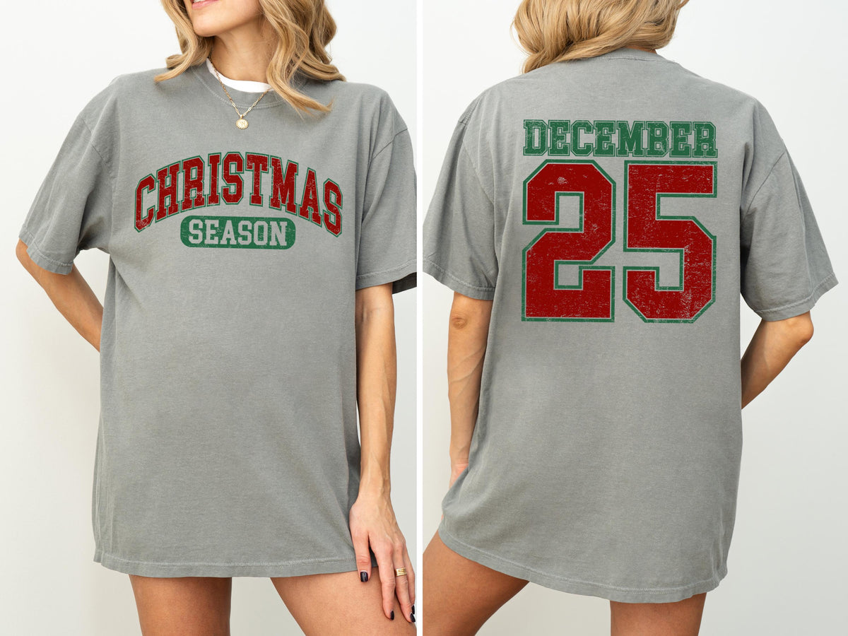 May include: A gray t-shirt with the words "CHRISTMAS SEASON" in red and green lettering on the front. The back of the shirt displays "DECEMBER 25" in red and green. The shirt has a vintage, distressed look.