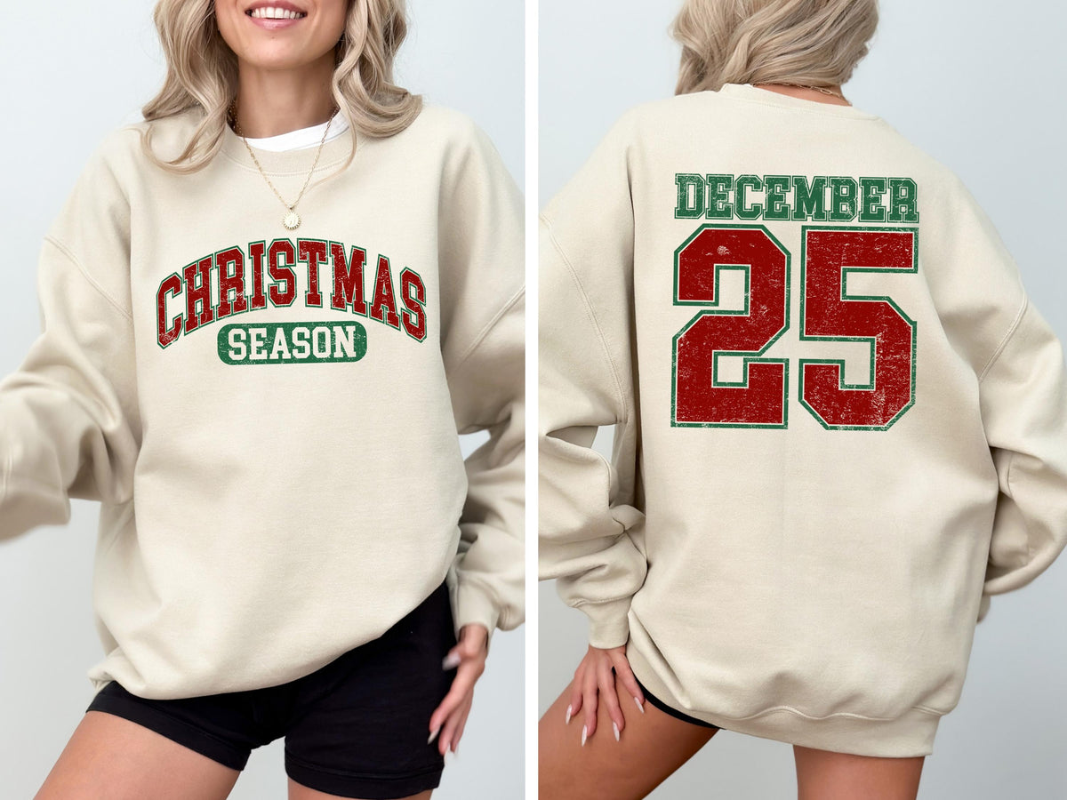 May include: A cream-colored sweatshirt with the words "CHRISTMAS SEASON" in red and green lettering on the front. The back of the sweatshirt displays "DECEMBER 25" in red and green. The sweatshirt is a festive holiday apparel item.