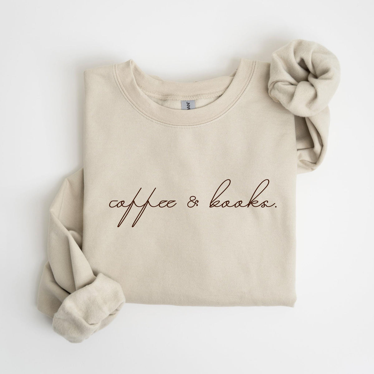 May include: Beige sweatshirt with the text "coffee & books" in a dark brown script. A matching scrunchie rests on the upper right. The sweatshirt has a crew neck and long sleeves, perfect for casual wear.