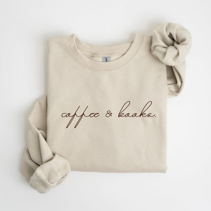 May include: Beige sweatshirt with the text "coffee & books" in a dark brown script. A matching scrunchie rests on the upper right. The sweatshirt has a crew neck and long sleeves, perfect for casual wear.