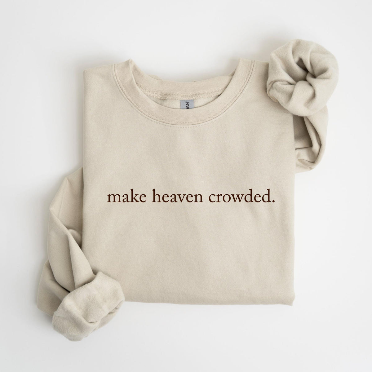 May include: A beige sweatshirt with the phrase "make heaven crowded" printed in a dark brown font. A matching beige scrunchie rests on the right side of the folded sweatshirt. The sweatshirt has long sleeves and a crew neck.