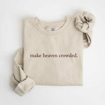 May include: A beige sweatshirt with the phrase "make heaven crowded" printed in a dark brown font. A matching beige scrunchie rests on the right side of the folded sweatshirt. The sweatshirt has long sleeves and a crew neck.