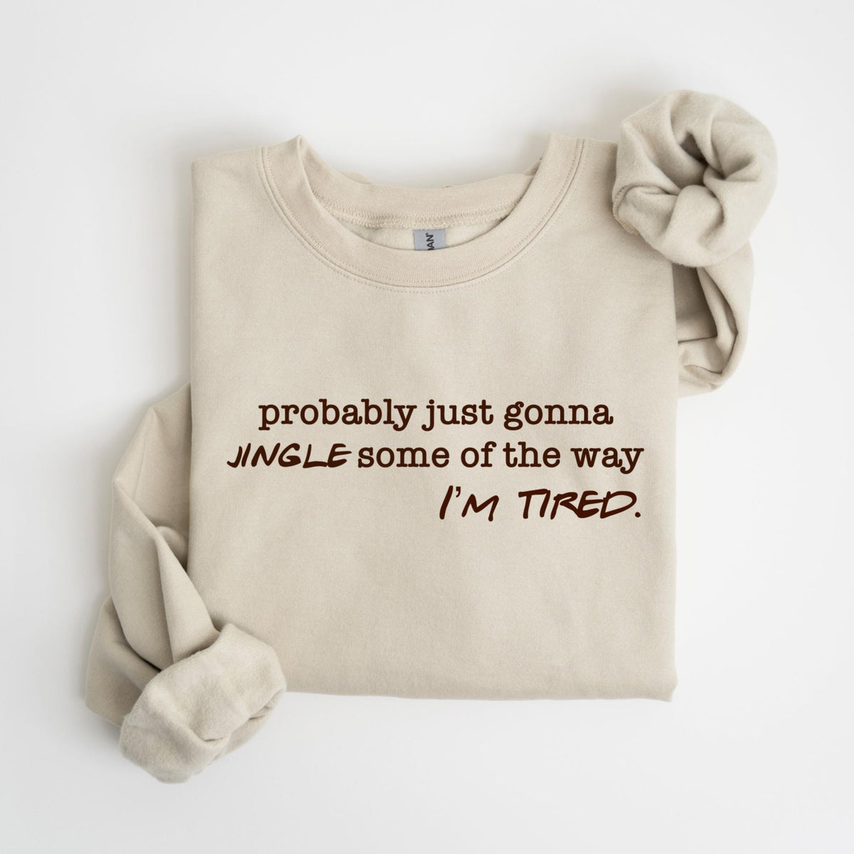 May include: A beige sweatshirt with the text "probably just gonna JINGLE some of the way I'M TIRED" in brown lettering. A matching scrunchie is next to the sweatshirt.