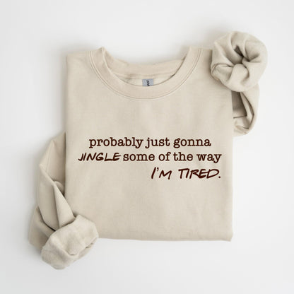 May include: A beige sweatshirt with the text "probably just gonna JINGLE some of the way I'M TIRED" in brown lettering. A matching scrunchie is next to the sweatshirt.