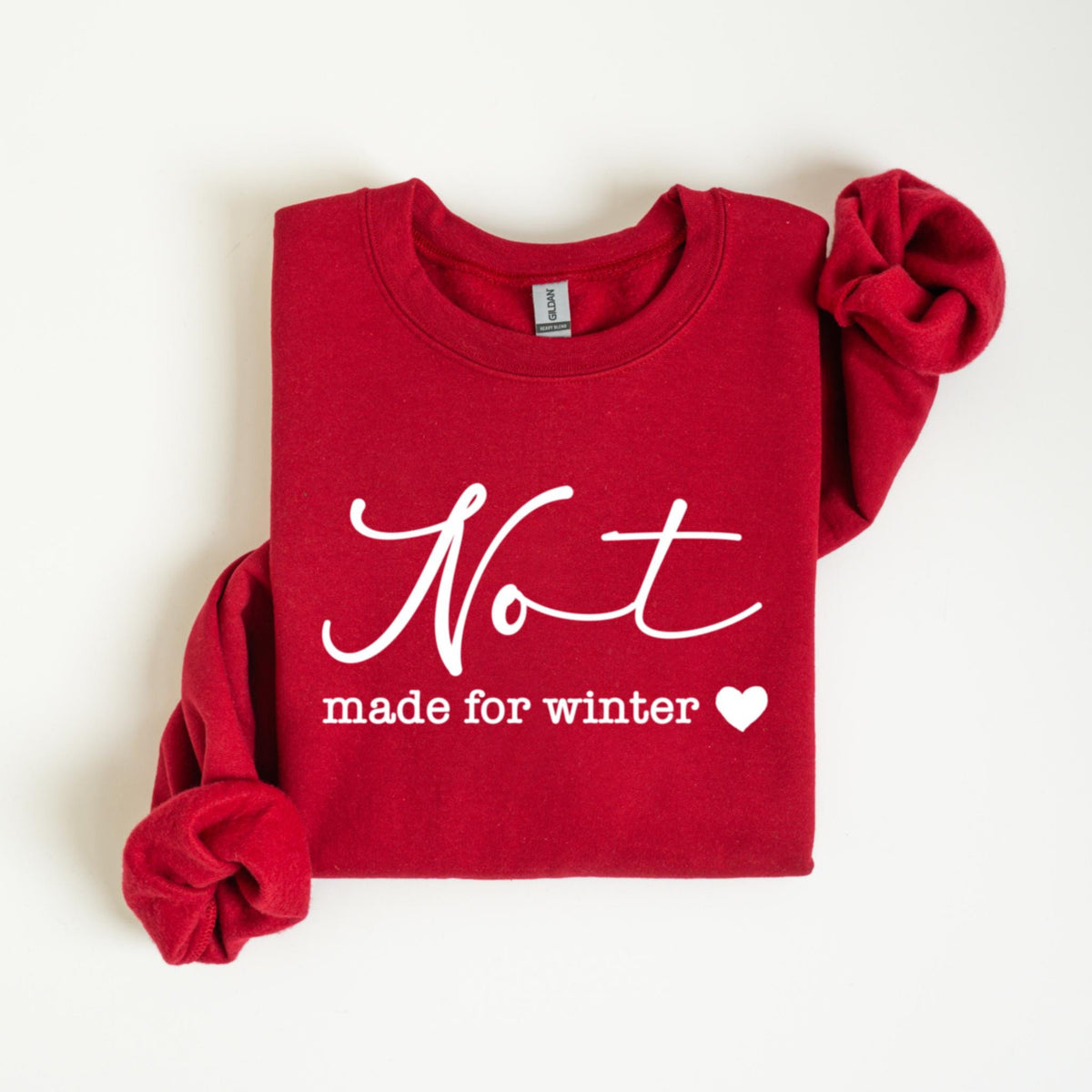 May include: A red sweatshirt with the word "Not" in a cursive font above the words "made for winter" and a white heart. The long sleeves are folded. The sweatshirt is made of a soft material.